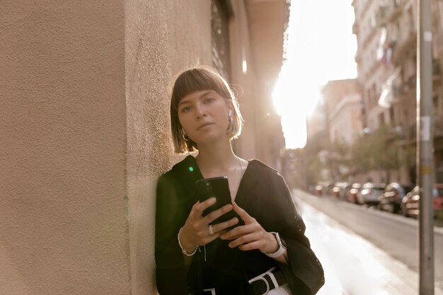 Primo piano foto esterna di una bella ragazza alla moda con acconciatura corta che indossa una camicia nera che tiene lo smartphone e guarda la fotocamera al sole