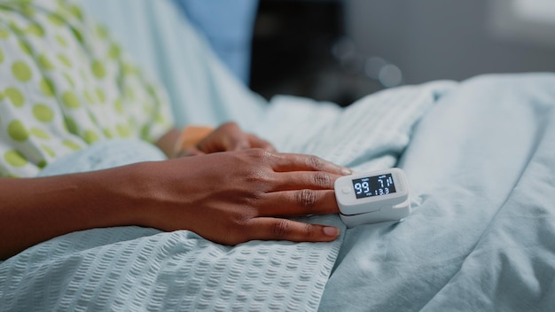 Close up of oximeter on finger of patient with illness in hospital ward bed. Sick woman having tool to measure oxygen saturation and pulse pressure to cure disease. Medical care