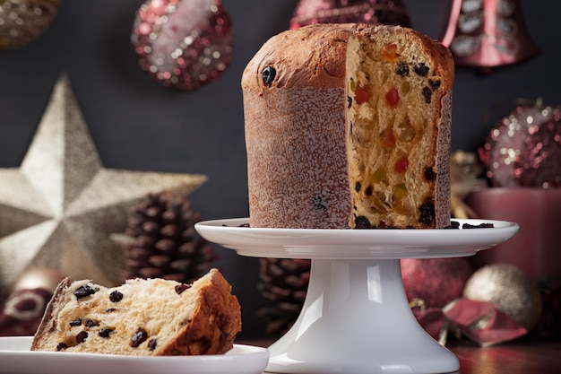 Close up of a panettone on a white plate with a slice served on the side, background with defocused and unsaturated christmas decoration.