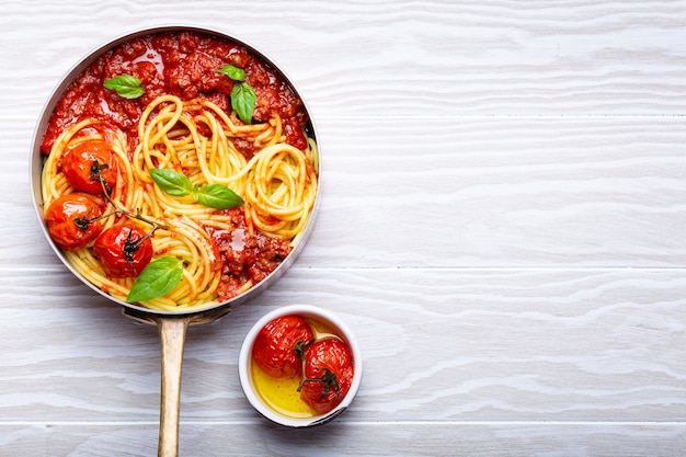 Primo piano di spaghetti di pasta con ragù di carne e pomodoro in padella servito con basilico e pomodorini su fondo rustico in legno bianco, vista dall'alto. cena tradizionale italiana, spazio per il testo