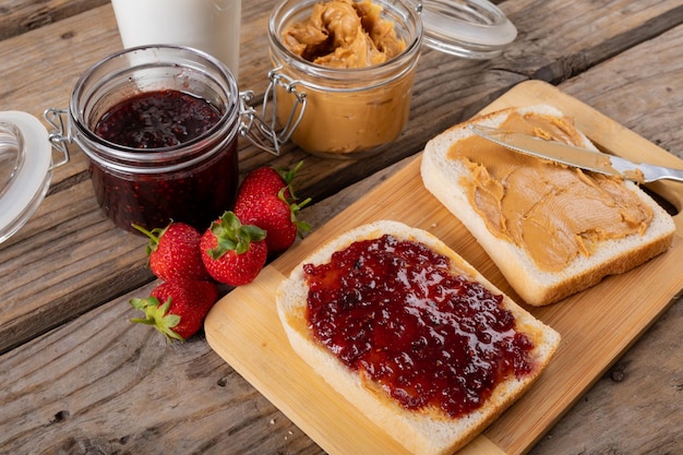 Photo close-up peanut butter and jelly sandwich on serving board with jars, strawberries and milk at table