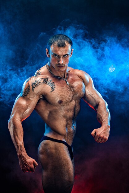 Close up on perfect abs. Strong bodybuilder with six pack.Strong bodybuilder man with perfect abs, shoulders,biceps, triceps and chest, personal fitness trainer flexing his muscles in blue, red smoke