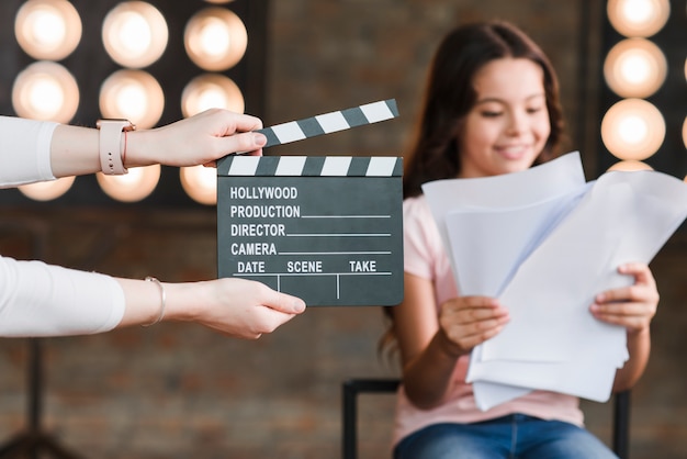 Close-up of a person holding clapper board in front of girl reading scripts