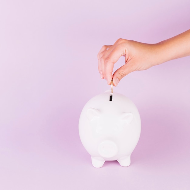 Photo close-up of a person inserting coin in white piggybank against pink background