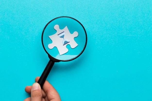 Close-up of a person's hand holding magnifying glass over hour glass icon on white puzzle over blue backdrop