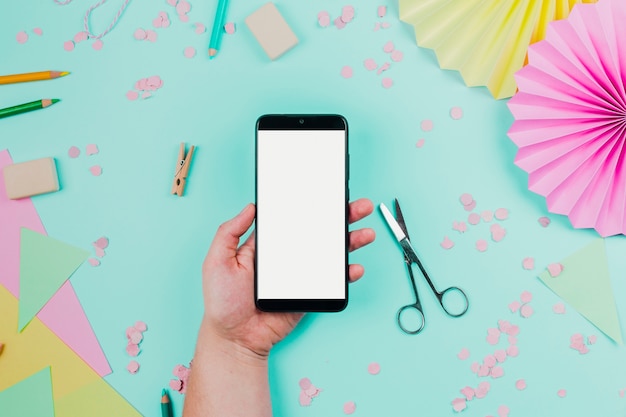 Close-up of a person's hand holding mobile phone with white screen display against teal backdrop