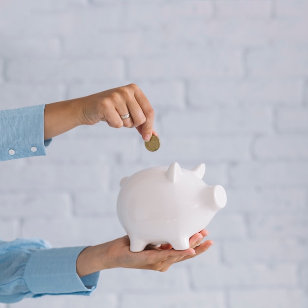 Close-up of a person's hand inserting coin in white piggybank