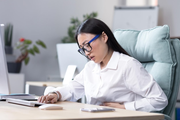 Primo piano foto dolore addominale sul posto di lavoro una giovane donna asiatica con gli occhiali lavora in ufficio a