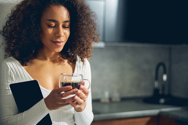 Close up photo of African American female standing in the kitchen and dreaming while holding coffee