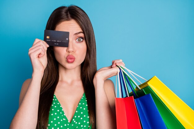 Close up photo of amazed girl close cover eye credit card make lips pouted plump hold many bags wear green dotted singlet crop tank-top isolated over blue color background