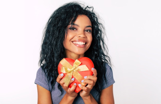 Close-up photo of amazing African ethnic girl with long black hair and sparkling eyes, who is over the moon while holding a red heart-shaped box with a golden ribbon in her hands