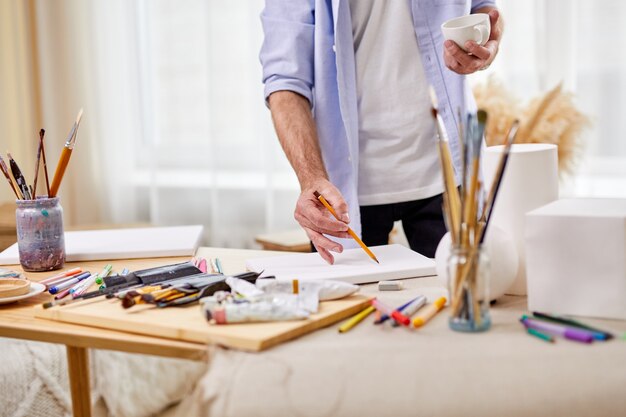 Close-up photo of artist man drawing with pencil on sheet, other different tools for painting on table