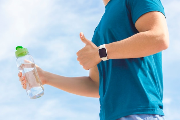 Close up photo of athlete's hand with smartwatch, bottle of water in hand. Unrecognizable person, fit man runner showing like gesture, thumb up. Healthy sport active fitness lifestyle, gadget concept