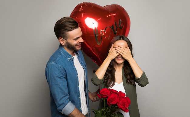 Close up photo of an attractive man, who is holding a red box in his hands, while his gorgeous girlfriend is smiling at him after receiving a bouquet of roses.