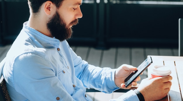 Close up photo of bearded businessman having a coffee break while surfing on mobile