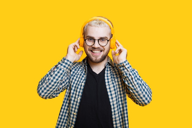 Close up photo of a bearded caucasian man with blonde hair wearing headphones and glasses posing on a yellow wall