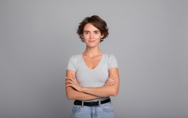 Close up photo of beautiful confident young girl with short hair and crossed arms wearing in tshirt looking on camera Studio shot of attractive female isolated against blank gray wall