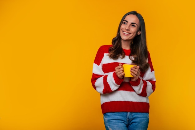 Close up photo of beautiful happy brunette woman with yellow cup of hot tea or coffee in hands isolated on yellow background