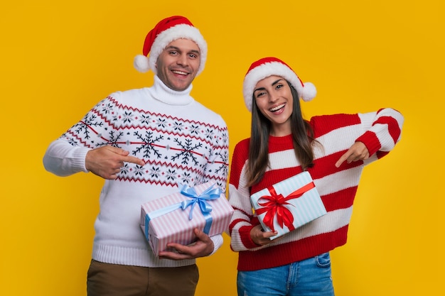 Close up photo of beautiful happy and excited young couple in love in Christmas clothes with gift boxes in hands while they pointing together on it.