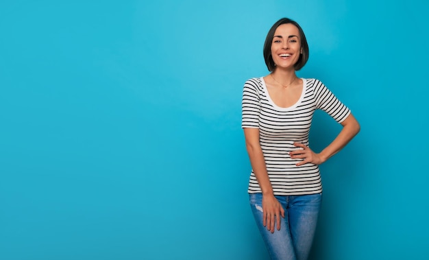 Close up photo of a beautiful smiling confident young brunette woman is posing in a striped t-shirt and isolated on blue background