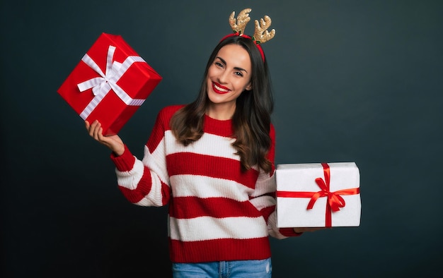 Крупным планом фото красивой улыбающейся модной женщины с рождественскими оленьими рогами на голове, когда она держит в руках подарочные коробки и веселится