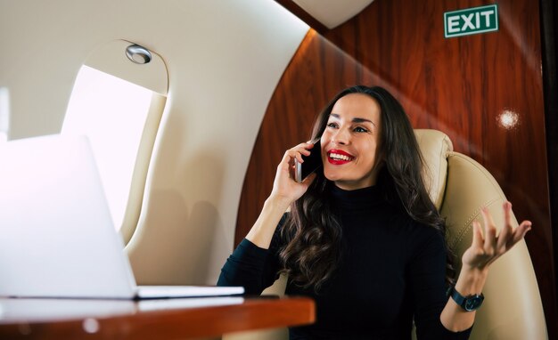 Foto ravvicinata di una bella donna in un abito casual, che sta parlando al telefono e bevendo caffè nero durante il suo volo in jet privato.