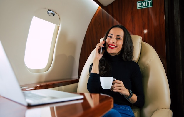 캐주얼 복장을 한 아름다운 여성이 전용기를 타고 비행하는 동안 전화 통화를 하고 블랙 커피를 마시는 클로즈업 사진.