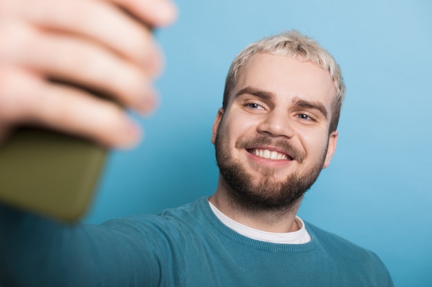 Close up photo of a blonde man with beard making a selfie using a phone on a blue studio wall