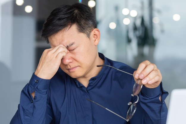 사무실에서 지친 젊은 아시아 남자가 손에 안경을 들고 앉아 직장에서 사진 번아웃을 닫습니다