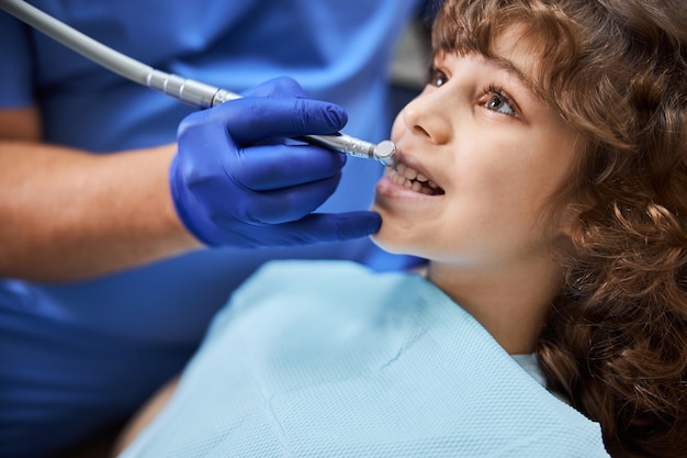 Close-up photo of a curly chestnut-haired child with dental drill near his teeth during a dental appointment