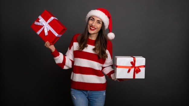 Close up photo of cute happy young woman in Santa hat with the bright colorful gift boxes in hands is having fun while posing isolated on gray background