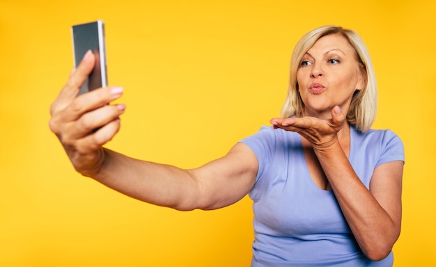 Close up photo of cute lovely blonde senior woman while she makes selfie photo on smart phone and sending air kiss