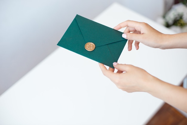 Close-up photo of female hands holding a green invitation envelope with a wax seal, a gift certificate, a postcard, a wedding invitation card.