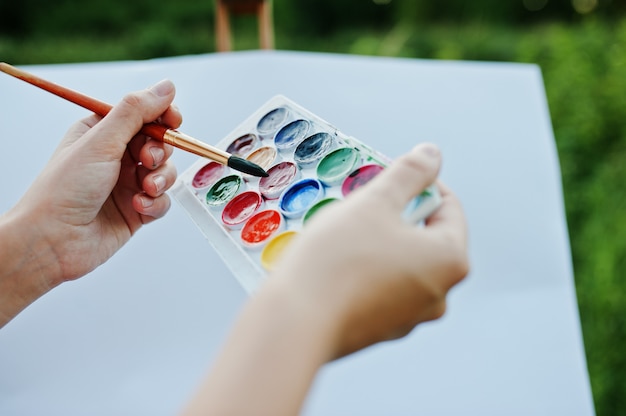 Foto foto del primo piano delle mani femminili che tengono le pitture dell'acquerello e una spazzola mentre dipingendo in natura.