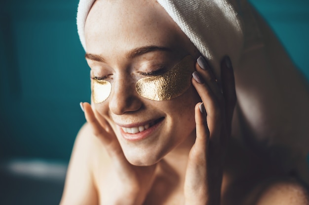 Close up photo of a freckles woman wearing golden eye patches covering her head with a towel and smile