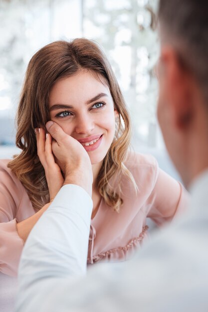 Close up photo of gentle woman looking on her man and taking pleasure when holding male hand at her face, while having date in restaurant