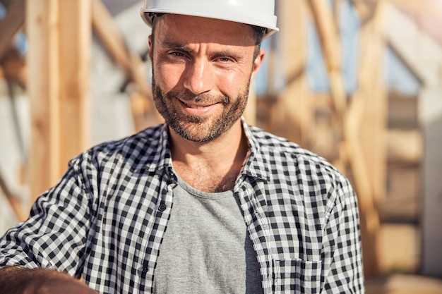 Close-up photo of a gladsome man in a hard hat and checked shirt looking at the camera