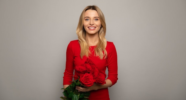 Close up photo of a gorgeous woman in a red outfit, who is posing in front, holding a bouquet of red roses and smiling with her eyes closed.