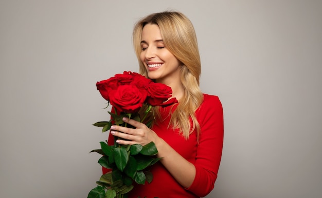 Close up photo of a gorgeous woman in a red outfit, who is posing in front, holding a bouquet of red roses and smiling with her eyes closed.