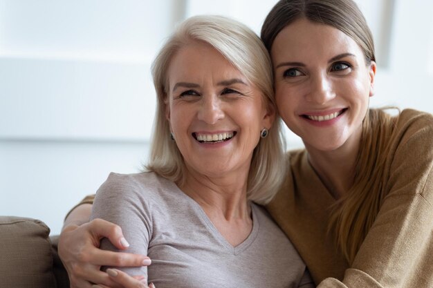 Close up photo grown up adult 30s daughter embracing elderly\
blonde mother diverse women sit on couch looking away feels happy\
enjoy moment spend time together showing care love and support\
concept