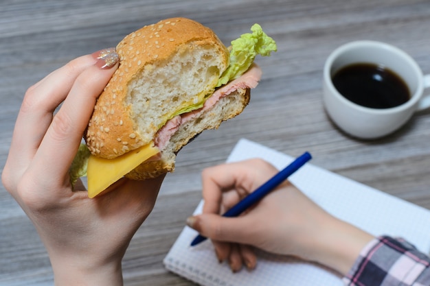 Close up photo of hands holding bitten burger and pen over copybook cup wooden table background