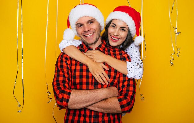 Foto foto ravvicinata di bella giovane coppia felice ed eccitata innamorata in cappelli di babbo natale in posa su uno sfondo giallo decorativo mentre celebra il natale e il capodanno