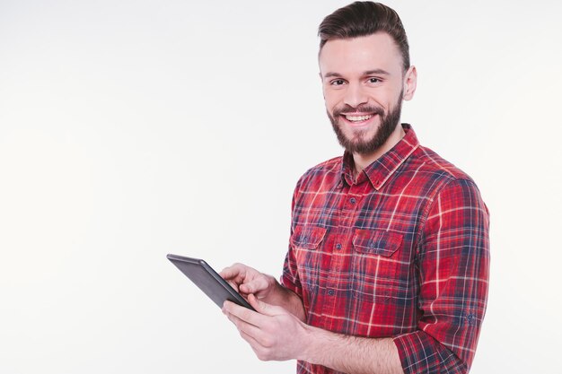 Close up photo of happy young bearded man in plaid shirt standing and using tablet isolated on white background