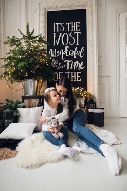 Фотография крупного плана маленькой девочки в вязаном свитере целует ее мать, сидя на диване на Рождество