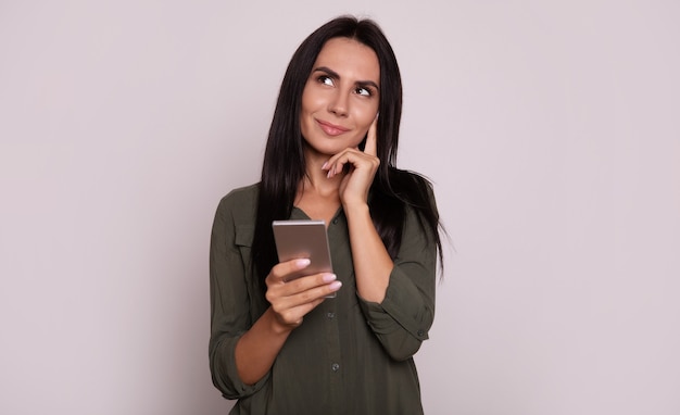 Close-up photo of a lovely woman in a green dress, who is posing with a sly smile, looking to the left upper corner and holding a smartphone in her right hand