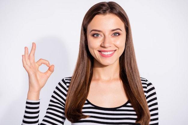 Chiuda sulla foto della donna adorabile che fa segno giusto che osserva con il sorriso a trentadue denti che indossa il ponticello a strisce isolato sopra la parete bianca