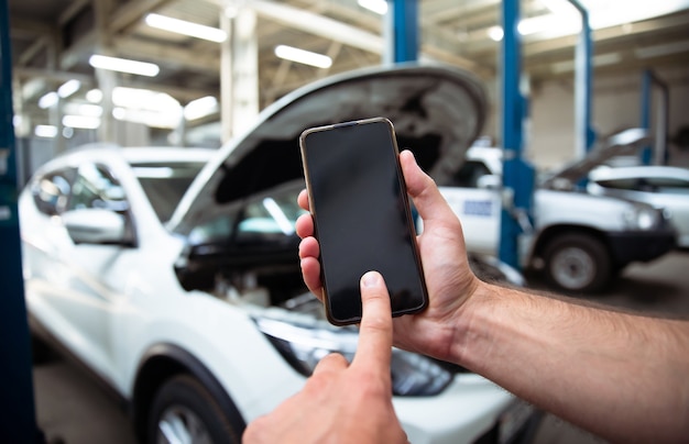 Close up photo of male hands with a blank screen of smartphone on background of auto service center with repairing cars