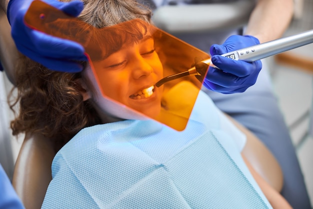 Фото Крупным планом - симпатичный мальчик, сидящий в стоматологическом кресле, лечится стоматологической лампой