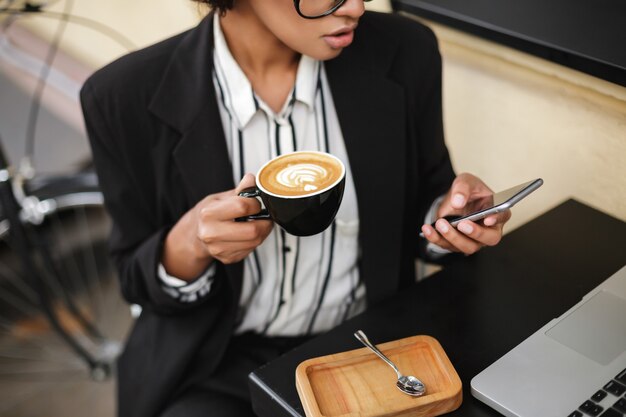 写真 携帯電話とコーヒーを片手にカフェのテーブルに座っているアフリカ系アメリカ人の女の子の写真を閉じる