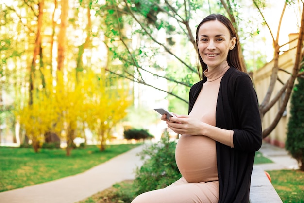 Foto ravvicinata di una donna incinta sorridente su una panchina nel parco mentre usa il suo smartphone